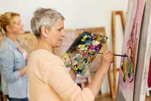 Senior woman in orange sweater painting a canvas on an easel and holding a pad with a lot of paint on it.